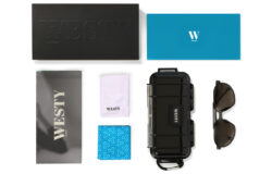 A collection of items including a black box labeled "WESTY," a blue box labeled "W," a silver envelope labeled "WESTY," a white cloth, a black protective case labeled "WESTY," a pair of black sunglasses, and a blue patterned cloth are all part of the King First Edition: Lunar Satin Titanium set.