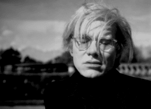 A black-and-white photograph of a person with platinum blonde hair and round glasses. They are wearing a high-collared dark sweater and are looking downward with a contemplative expression. The background is slightly blurred, featuring trees and a distant structure.