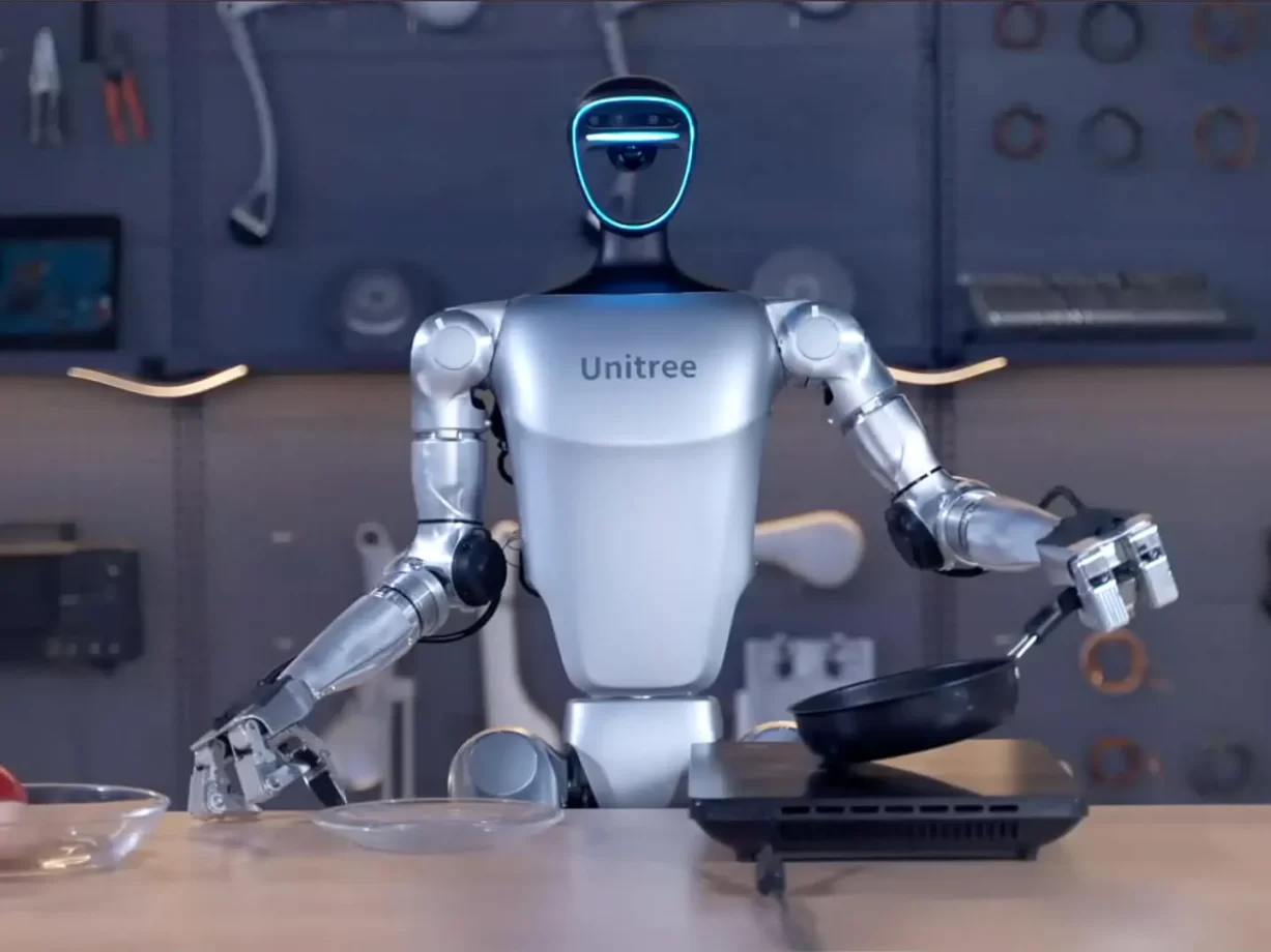 A humanoid robot with "Unitree" on its chest is standing in a kitchen-like setting, holding a frying pan in one hand and a spatula in the other. It appears to be cooking, with various tools visible in the background.