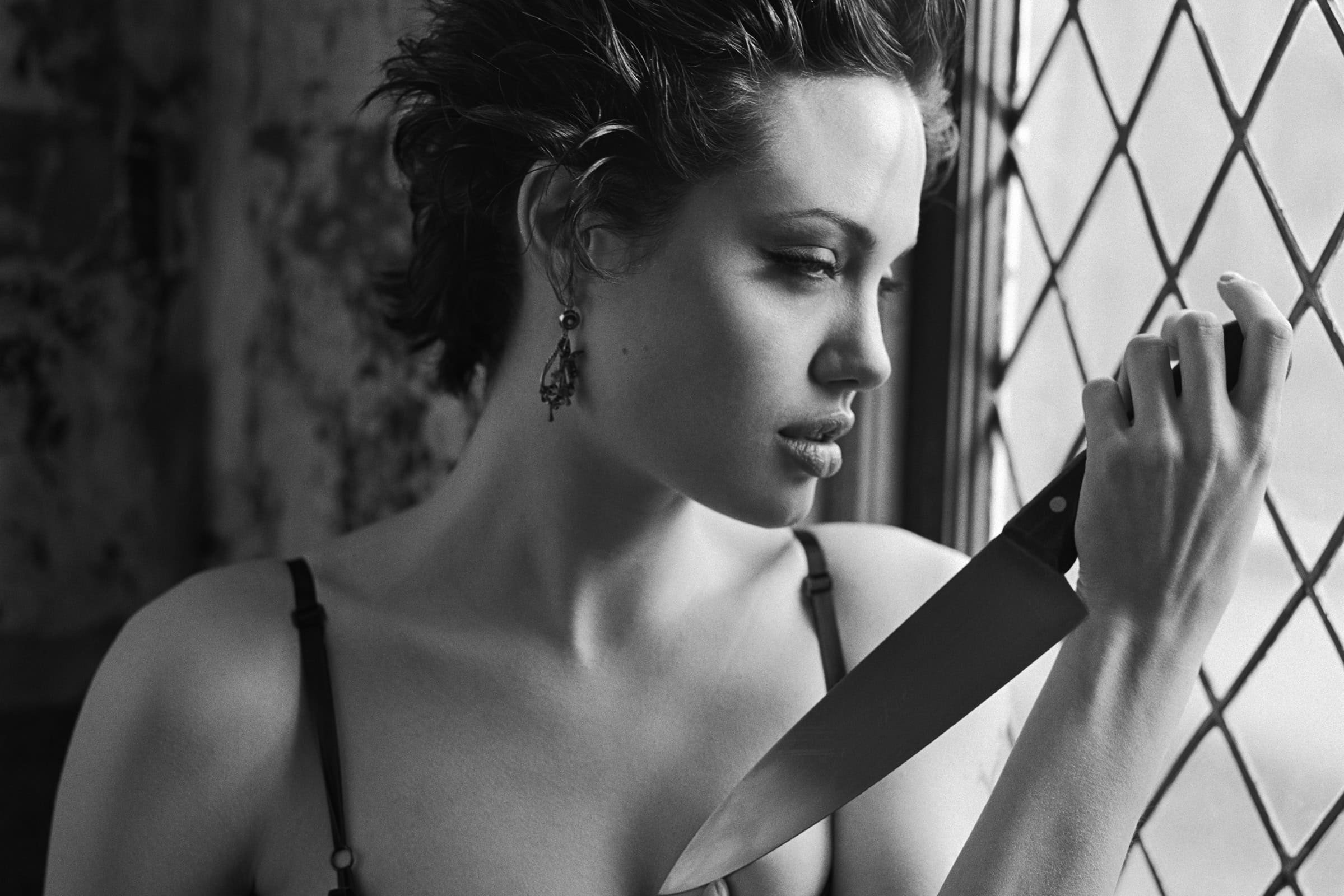 A black and white photo of a person holding a knife thoughtfully by a window with diamond-patterned panes. They have short, styled hair and are wearing earrings. The lighting creates a dramatic, introspective mood.