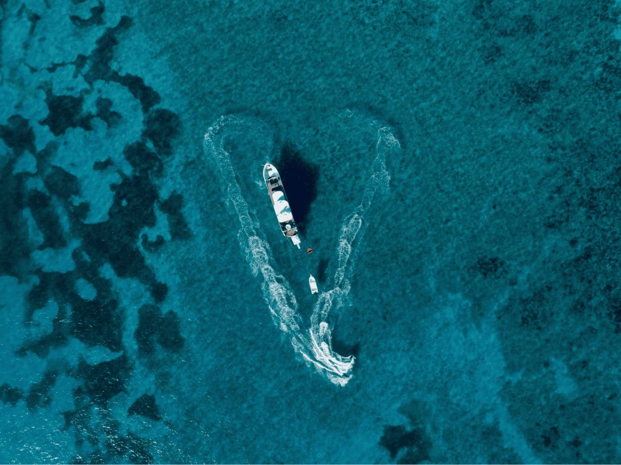 Aerial view of a boat on clear blue water, creating a heart-shaped wake with its path. A small boat follows behind, and the surrounding water shows varying shades of blue and patches of dark coral or rocks.