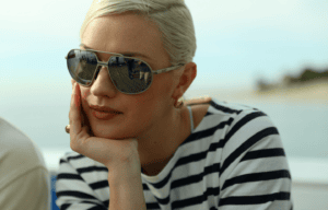 A woman with blonde hair, wearing sunglasses and gold earrings, rests her chin on her hand. She is dressed in a black and white striped shirt and is outdoors, with blurred beach scenery in the background.